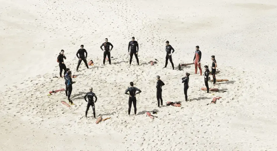 A large group of people standing in a circle on a beach. Each person is wearing a wet suit and standing next to a rescue tube